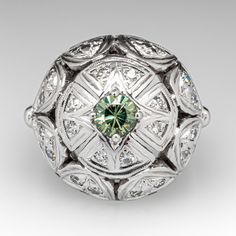 This vintage ring features a domed gallery with a pierced design that is accented with one (1) prong set, round modified brilliant natural Demantoid garnet and twenty-four (24) bead set, round single cut diamonds. The ring measures 17.8mm at the top, rises 9.3mm above the finger, tapering to 1.8mm wide and 1.1mm thick at the base of the shank. It is crafted in 14k white gold and is currently a size 5.25. Several diamonds have light unnoticeable chips. Vintage Green Diamond Platinum Ring, Vintage Green Diamond Ring In Platinum, Art Deco Round Emerald Ring With Center Stone, Elegant Domed Emerald Ring, Heirloom Style Green Diamond Cluster Ring, Heirloom Green Diamond Cluster Ring, Antique Emerald Ring In White Gold, Antique White Gold Emerald Ring, Vintage Green Diamond Ring With Center Stone