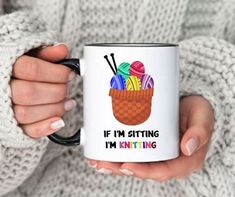 a woman is holding a coffee mug with knitting items in it and the words if i'm sitting, i'm knitting