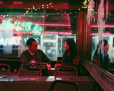 two people sitting at a table in a restaurant with neon lights on the wall behind them