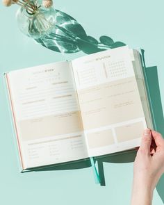 a person holding an open book on top of a table next to a vase with flowers