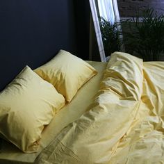 a bed with yellow sheets and pillows in front of a window next to a potted plant