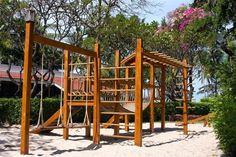 a wooden swing set in the sand near some bushes and trees with pink flowers behind it