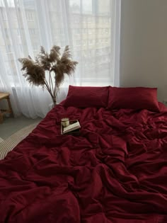 an unmade bed with red sheets and pillows in front of a window, next to a plant