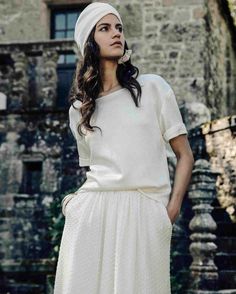 a woman wearing a white hat and dress standing in front of a stone building with her hands on her hips