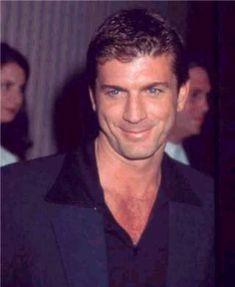 a man in a black shirt and blazer smiling at the camera with other people behind him