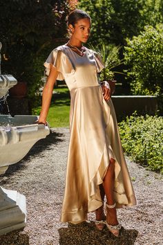 a woman standing in front of a fountain wearing a gold dress and high heeled sandals