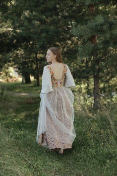 a woman in a long dress is walking through the grass with trees and bushes behind her