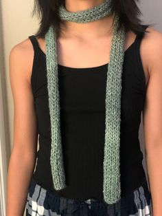 a woman wearing a green knitted scarf and black tank top standing in front of a white wall