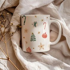 a white coffee mug with christmas decorations on it sitting on a bed next to dried flowers