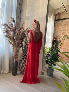 a woman in a red dress standing next to a plant and looking out the window