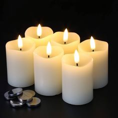 six white candles with one lit and the other on top of some silver coins in front of them