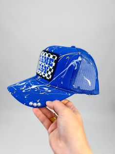 Score style points with this Game Day Checkered Splatter Paint Cap! The unique splatter design and checkered patch add a fun twist to a classic trucker hat. Perfect for showing your support for the blue team on game day. Key Features Adjustable snapback Lightweight + Comfortable Checkered Game Day Patch Splatter paint Pearl details Blue Snapback Trucker Hat With Letter Print, Blue Snapback Hat With Letter Print And Curved Bill, Blue Flat Bill Baseball Cap For Game Day, Blue Trucker Snapback Hat With Letter Print, Blue 5-panel Trucker Hat For Baseball Season, Blue Letter Print Snapback Hat For Streetwear, Blue Adjustable Trucker Hat Hip Hop Style, Blue Adjustable Hip Hop Trucker Hat, Blue Baseball Cap For Sports Fans With Flat Bill