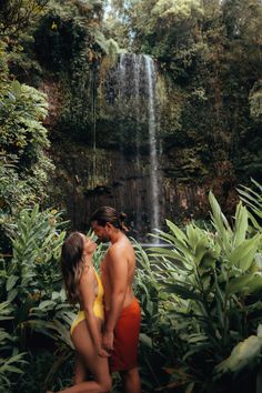 a man and woman in swimsuits standing next to a waterfall with greenery