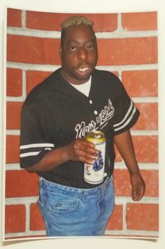 a man holding a can of beer standing in front of a brick wall with his hands on his hips