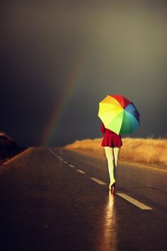 a woman walking down the road with an umbrella over her head and a rainbow in the background