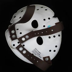 a white mask with brown leather straps and holes on the face is shown in front of a black background