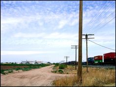 a dirt road with power lines, telephone poles and train cars on the tracks in the distance