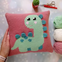 a crocheted dinosaur pillow sitting on top of a table next to balls of yarn