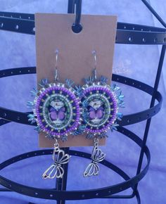 There is a lot going on here. The centers are resin ovals with a simple purple butterfly in it, surrounded by purple and green beads that give it a foresty feel. And a butterfly charm that is just as large as the earring itself. Colorful dragon scale beads are used on the edge. Its a lot and its wonderful. Bohemian Embellished Beaded Earrings, Purple Bohemian Beaded Jewelry, Purple Beaded Bohemian Jewelry, Handmade Purple Beaded Earrings, Purple Bohemian Beads For Festival, Bohemian Lavender Beaded Dangle Earrings, Handmade Adjustable Lavender Beaded Earrings, Handmade Purple Bohemian Beads, Purple Handmade Beads For Crafting