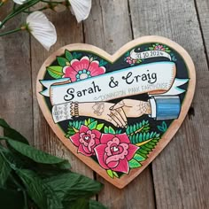 a wooden heart shaped sign with flowers on the side and two hands holding each other