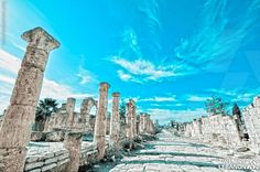 the ancient ruins are surrounded by stone pillars and palm trees, under a blue sky with wispy clouds