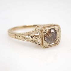 a fancy ring with a brown diamond in the center on a white surface, it is gold and has filigree details