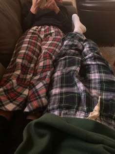 a man laying on top of a couch next to a woman in plaid pajamas and holding a cell phone