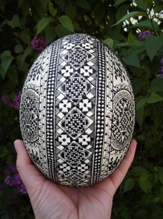 a hand holding an ornately decorated egg in front of some purple and white flowers