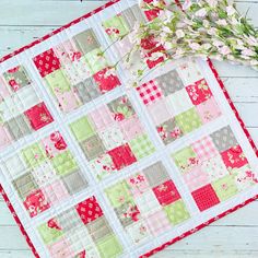 a close up of a quilt on a table with flowers next to it and a plant in the background