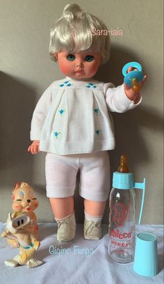 a doll with blonde hair and blue eyes is holding a toy in her hand while standing next to a bottle