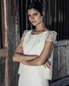 a woman standing with her arms crossed in front of a wooden wall and gate, wearing a white dress