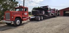 a tractor trailer hauling a dump truck down a dirt road next to a red barn