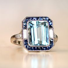 A stunning gemstone ring that centers on a 3.20 carat natural emerald-cut aquamarine. Baguette-cut diamonds and natural French-cut sapphires surround the aquamarine. Additional diamonds line the shoulders. This ring is platinum and displays an open-work under-gallery. ✦ GEMSTONE SPECIFICATIONS: Center Stone: Aquamarine Gemstone Weight: 3.20 Carats Gemstone Cut: Emerald Cut ✦ ENGAGEMENT RING SPECIFICATIONS: Stones: Aquamarine, Diamonds, Sapphires Material: Platinum Ring Style: Art Deco ✦ WHAT COM Emerald Shape Engagement Rings, Diamond Ring With Halo, Ring With Halo, Estate Diamond Jewelry, Rings To Make, Emerald Cut Rings, Ring Inspiration