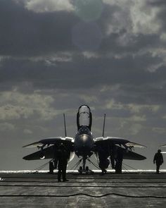 a fighter jet sitting on top of an airport tarmac under a cloudy sky with two men standing next to it