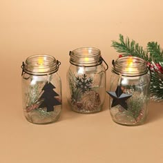 three glass jars with christmas decorations and candles in them on a brown surface next to a pine tree