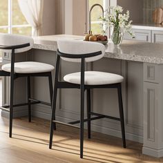 two white stools sitting in front of a kitchen counter with flowers on it and an open window