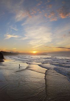 a person walking on the beach at sunset with waves coming in from the water and clouds