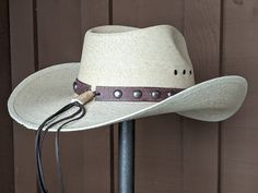 Chet starts with a Mexican soft palm leaf hat body that has a 3 3/4" brim and 5 1/4" crown. The band is simple with distressed brown leather and antique nickel color spots. The back of the band has a deer antler bead for accent. A neutral feather combo completes the hat. Note: Please allow 3-4 weeks. Rustic Adjustable Fedora With Flat Crown, Natural Hat Band With Flat Brim For Country Events, Adjustable Natural Hat Band For Rodeo, Rustic Adjustable Hat With Curved Brim, Rustic Adjustable Brimmed Hat, Rustic Adjustable Short Brim Hat, Natural Hat Bands For Kentucky Derby, Natural Color Adjustable Western Hat Bands, Adjustable Natural Color Western Hat Band