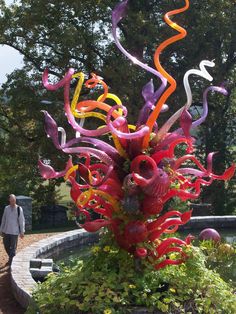 a colorful sculpture is in the middle of a flower bed with people walking around it