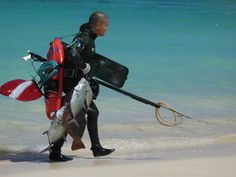 a man walking on the beach with a large fish in his hand and luggage strapped to his back