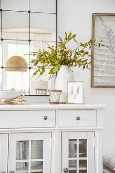 a white dresser topped with a vase filled with flowers and greenery next to a mirror