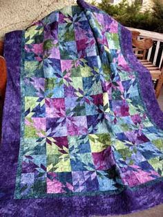 a purple and green quilt sitting on top of a wooden bench next to a tree