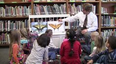 a man standing in front of a group of children sitting on the floor next to bookshelves