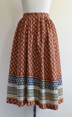 "This gorgeous skirt is gently gathered at the waist and comes in a handsome shade of rust--somewhere at the nexus of brown, red, and orange. The print features golden yellow paisley-like motifs as well as ornate bands of white and navy blue with pale green accents. Zippered back closure (nylon coil teeth) with an attractive plastic button. Good, wearable vintage condition. One tiny hole on the front left about the size of a pinhead (pictured), which appears to be a burn mark. We spotted two or Vintage Brown Flowy Skirt, Bohemian Brown Pleated Maxi Skirt, Traditional Long Brown Skirt, Brown Vintage Tiered Skirt, Vintage Brown Tiered Skirt, Vintage Cotton Brown Skirt, Vintage Long Brown Skirt, Vintage Skirt With Elastic Waistband In Relaxed Fit, Vintage Relaxed Skirt With Elastic Waistband