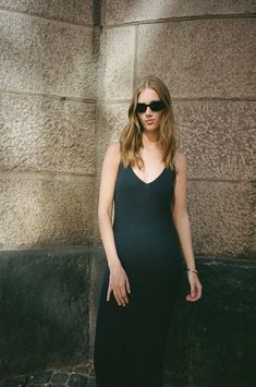 a woman standing in front of a wall wearing sunglasses and a black dress with her hands on her hips