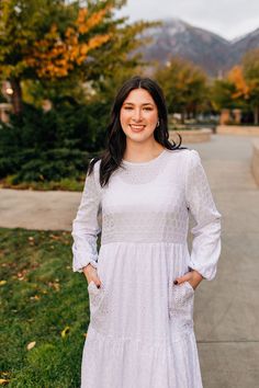 a woman standing on the sidewalk wearing a white dress and smiling at the camera with mountains in the background