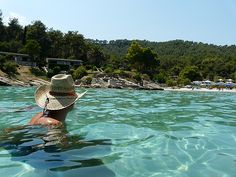 a woman wearing a straw hat is swimming in clear blue water near the beach and trees