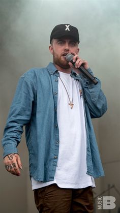 a man standing on stage with a microphone in his hand and wearing a baseball cap