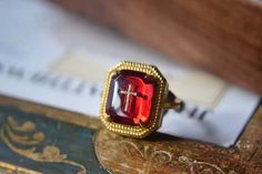 The Cross Faith Signet Ring is a bold and symbolic statement piece that blends vintage elegance with spiritual meaning. Featuring a deep ruby-red crystal with an embedded golden cross, this ring serves as a reminder of inner strength and unwavering faith. The octagonal gold setting, adorned with intricate beading, frames the gem beautifully, adding a regal touch to this unique design. Perfect for those who wish to wear their beliefs with pride, the Cross Faith Signet Ring is more than just an accessory—it's a reflection of personal conviction and style. Details Cross Faith Signet Ring in Red Gold pleated Adjustable opening Missy Jewelry Dragon Breath Collection Luxury Heirloom Red Signet Ring, Elegant Red 14k Gold Signet Ring, Luxury Red Signet Ring With Polished Finish, Collectible Red Gemstone Signet Ring, Antique Red Gemstone Signet Ring, Dragon Breath, Golden Cross, Intricate Beading, Unwavering Faith