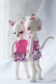 two white cats standing next to each other on top of a white surface with one cat wearing a pink dress